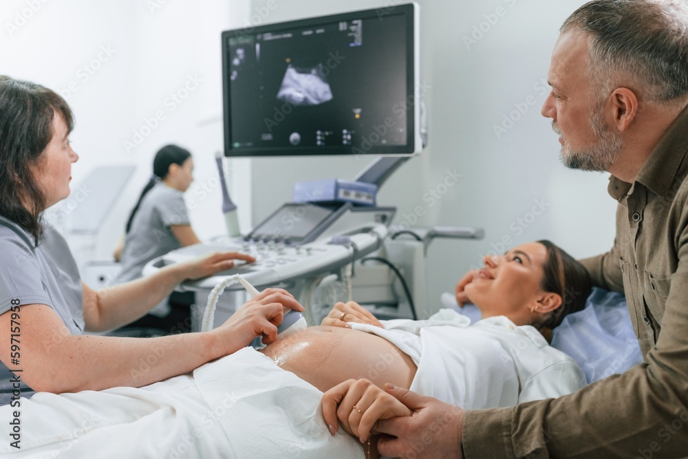 Happy husband is sitting and supporting his wife. Pregnant woman is lying down in the hospital, doctor does ultrasound Happy husband is sitting and supporting his wife. Pregnant woman is lying down in the hospital, doctor does ultrasound