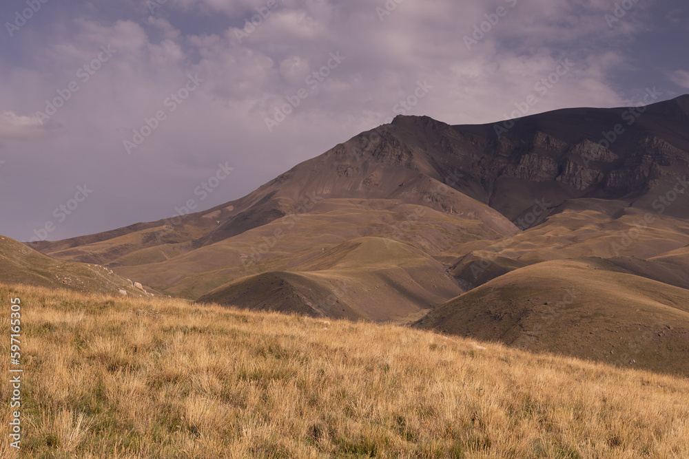 Mountains landscape - dark mountain ridges with hilly slopes in sunny autumn day with white fluffy clouds and golden sunbeams, yellow spikelets of dried plants on meadow. Majestic journey in Dagestan.