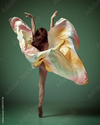 Fototapeta Naklejka Na Ścianę i Meble -  Back view of one charming ballerina, ballet dancer wearing fancy colorful dress posing on tiptoe over dark green studio background. Aesthetic of movement