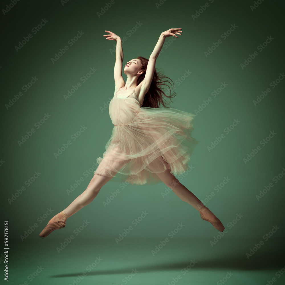 Young and incredibly beautiful ballerina wearing tulle dress jumping gracefully over dark green studio background. Demonstrate flexibility - obrazy, fototapety, plakaty 