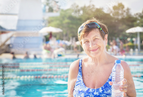Active healthy senior (elderly) woman (over the age of 50) in sport goggles and with towel and bottle of water smiling with happy face after swimming in summer day. Swimming pool background.