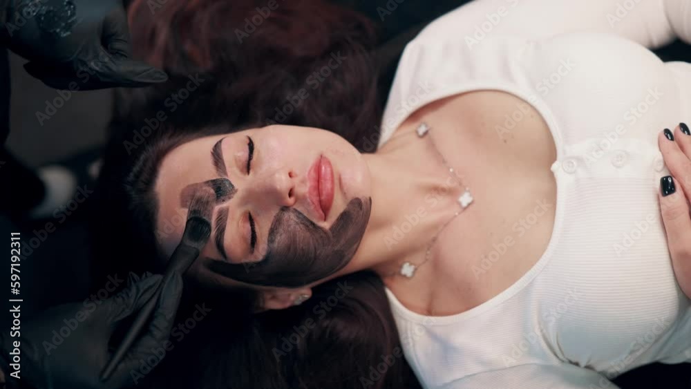 Cosmetologist applying black mask on pretty woman face. Female in spa having facial procedures. Top view. Cosmetologist applying black mask on pretty woman face. Female in spa having facial procedures. Top view.