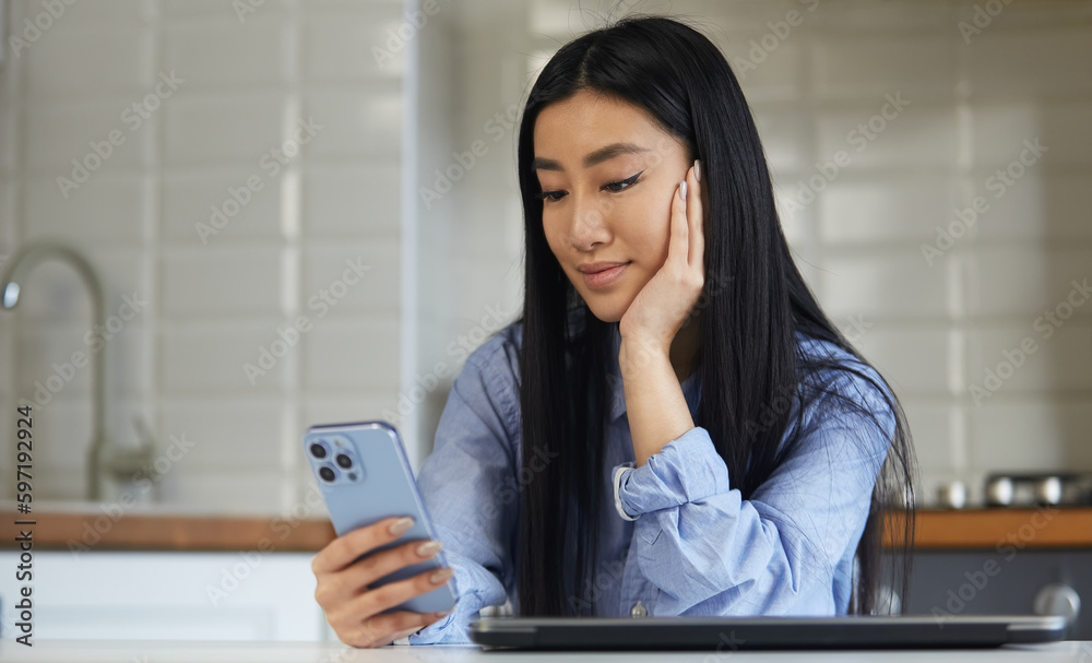 Cheerful young Asian girl browsing mobile app with a smile. Beautiful POC female using modern blue smartphone in a home kitchen Cheerful young Asian girl browsing mobile app with a smile. Beautiful POC female using modern blue smartphone in a home kitchen