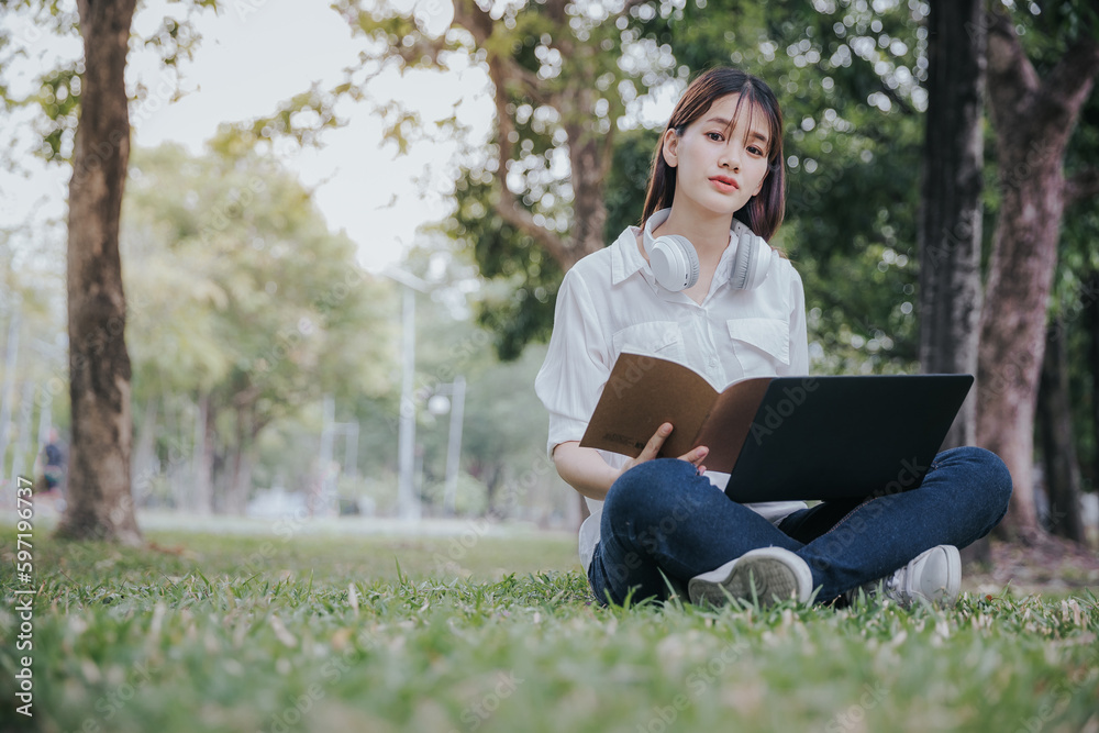 Portrait of young asian woman in white shirt sitting on green grass in park with legs crossed during summer day while using laptop and wireless headphones for video call. Portrait of young asian woman in white shirt sitting on green grass in park with legs crossed during summer day while using laptop and wireless headphones for video call.
