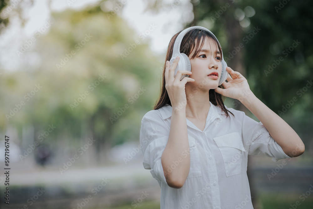 Portrait of young asian woman in white shirt sitting on green grass in park with legs crossed during summer day while using laptop and wireless headphones for video call. Portrait of young asian woman in white shirt sitting on green grass in park with legs crossed during summer day while using laptop and wireless headphones for video call.