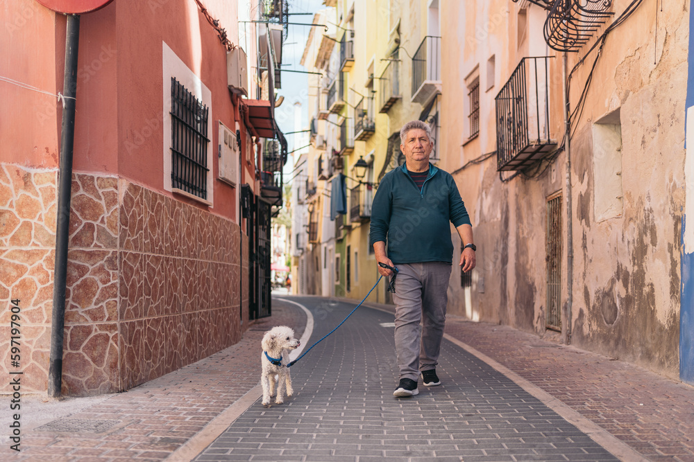 Front view image of gray-haired gentleman walking his white curly-haired dog.