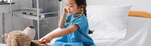 asian girl examining teddy bear with toy stethoscope on hospital bed, banner.