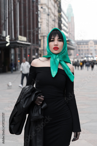 A woman with a perfect figure and red lips in a short black dress, a bright scarf on her head. Posing in the city