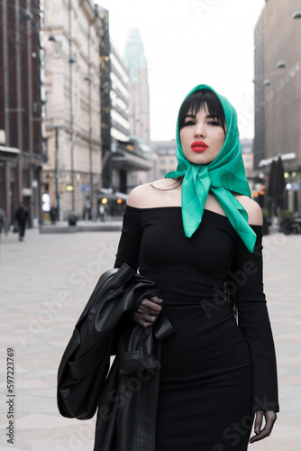 A woman with a perfect figure and red lips in a short black dress, a bright scarf on her head. Posing in the city