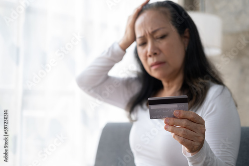 Unhappy Asian senior mature woman holding her credit card, feeling headache and stressed, having financial problems.