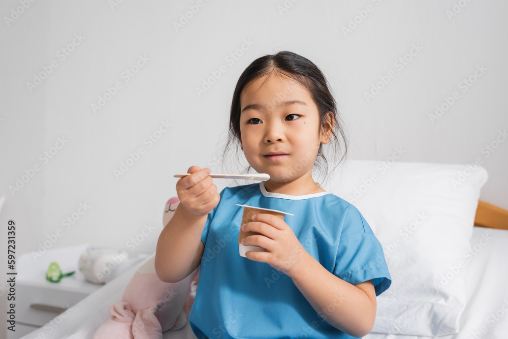 positive asian child in hospital gown eating delicious yogurt in clinic.