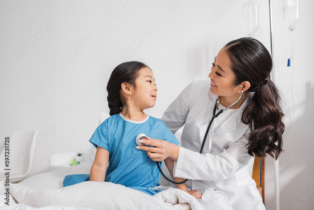 positive doctor in white coat examining asian girl with stethoscope in hospital ward. positive doctor in white coat examining asian girl with stethoscope in hospital ward.