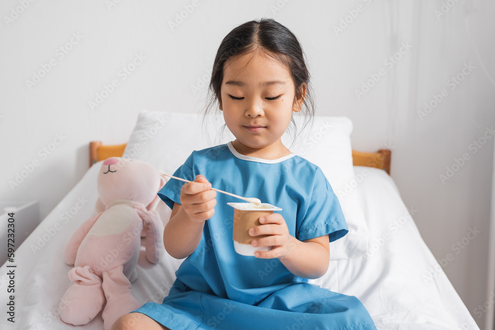 little asian girl in hospital gown eating delicious yogurt during breakfast in clinic. little asian girl in hospital gown eating delicious yogurt during breakfast in clinic.