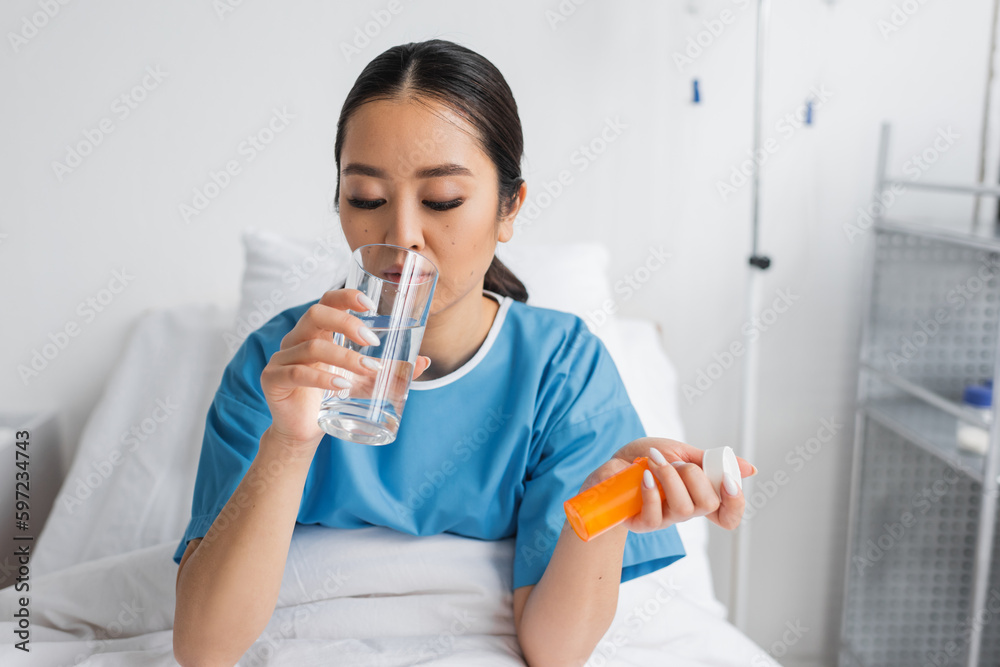 young asian woman drinking water while holding pills container on bed in clinic. young asian woman drinking water while holding pills container on bed in clinic.