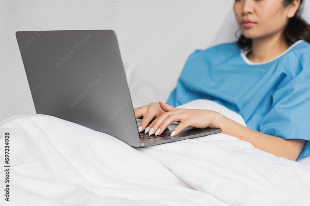 partial view of asian woman typing on laptop on bed in hospital ward. partial view of asian woman typing on laptop on bed in hospital ward.