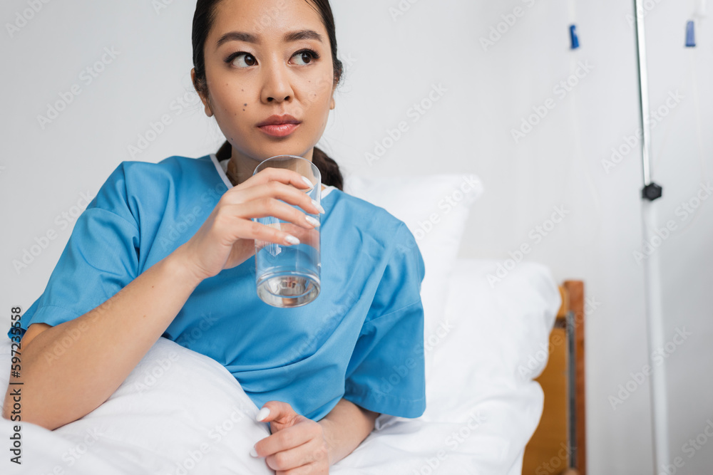 thoughtful asian woman holding glass of water and looking away in hospital ward. thoughtful asian woman holding glass of water and looking away in hospital ward.