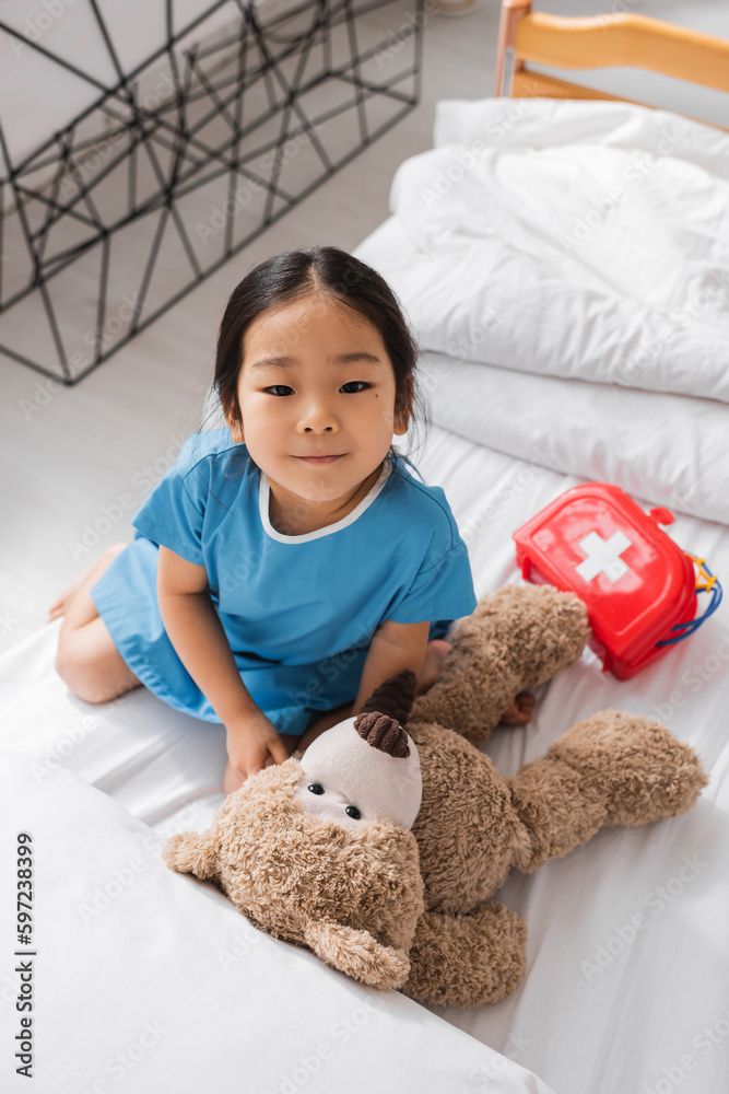 top view of asian girl smiling at camera while playing with teddy bear and toy medical equipment on hospital bed. top view of asian girl smiling at camera while playing with teddy bear and toy medical equipment on hospital bed.