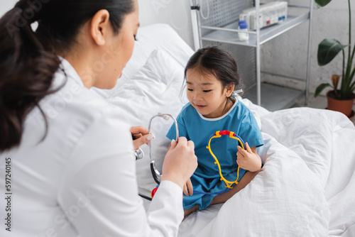 blurred doctor showing stethoscope to positive asian girl sitting with toy on hospital bed.