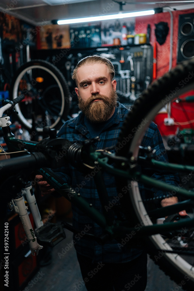 Obraz premium Vertical portrait of brutal cycling repairman standing by bicycle in repair workshop with dark interior, looking at camera. Concept of professional repair and maintenance of bicycle transport.
