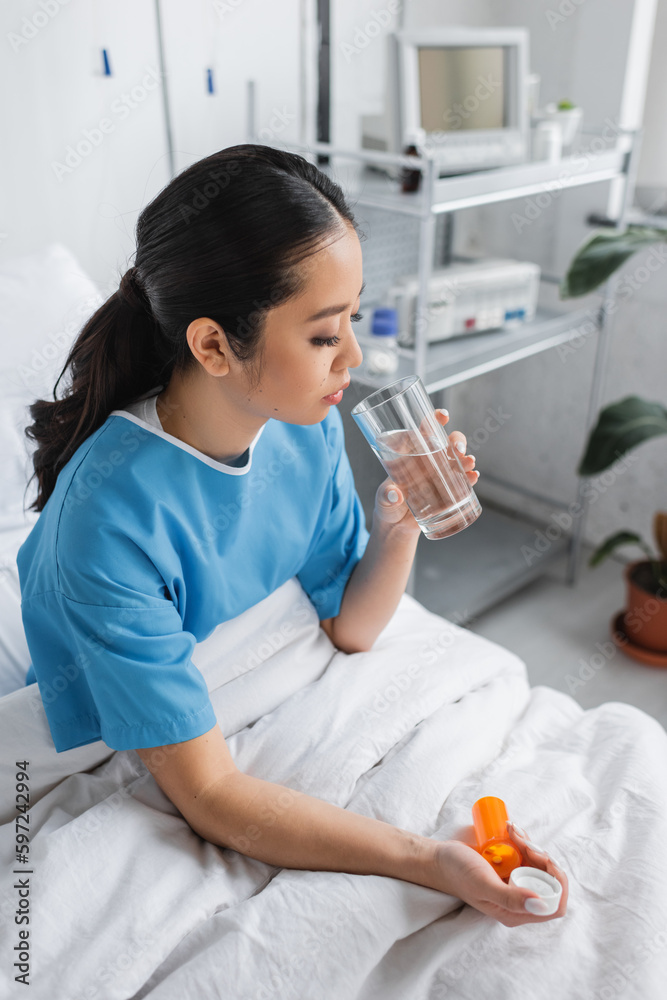 brunette asian woman in hospital gown sitting on bed with glass of water and pills container. brunette asian woman in hospital gown sitting on bed with glass of water and pills container.
