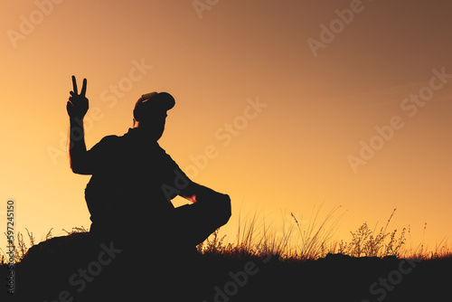 silhouette of a man in a cap sitting outside on the grass, two fingers up