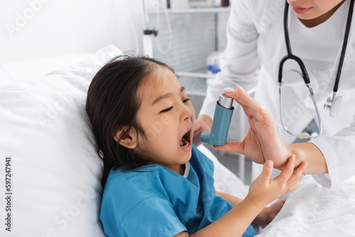 asian girl opening mouth near doctor using inhaler in pediatric clinic.