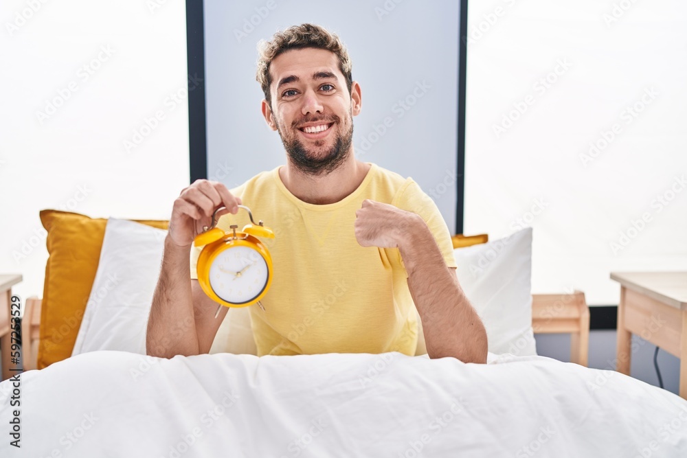 Hispanic man with beard holding alarm clock in the bed pointing finger to one self smiling happy and proud Hispanic man with beard holding alarm clock in the bed pointing finger to one self smiling happy and proud