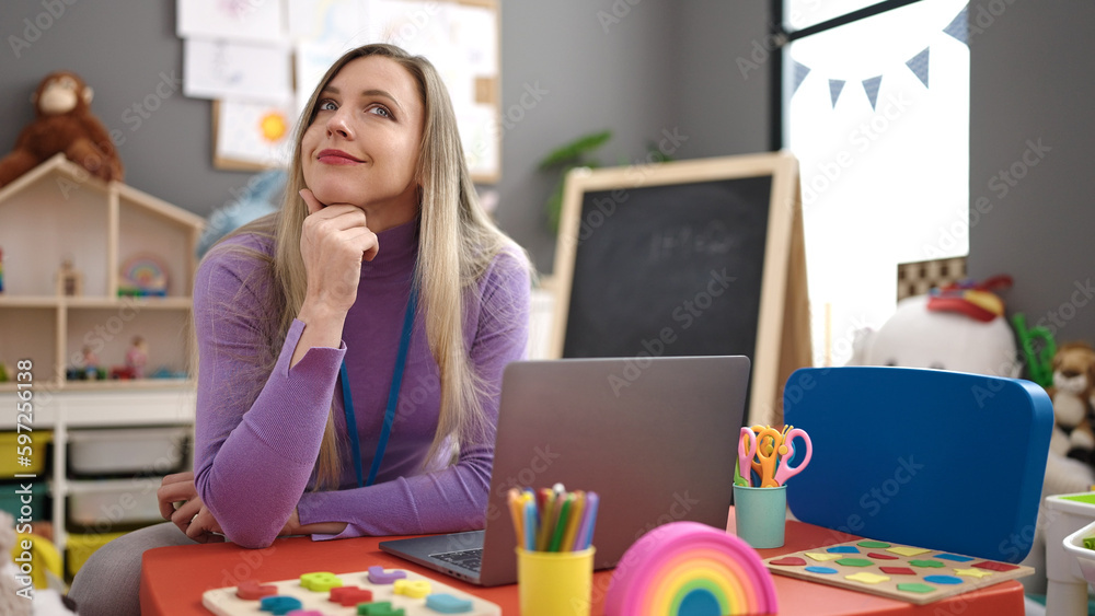 Young blonde woman preschool teacher using laptop sitting on table at kindergarten Young blonde woman preschool teacher using laptop sitting on table at kindergarten