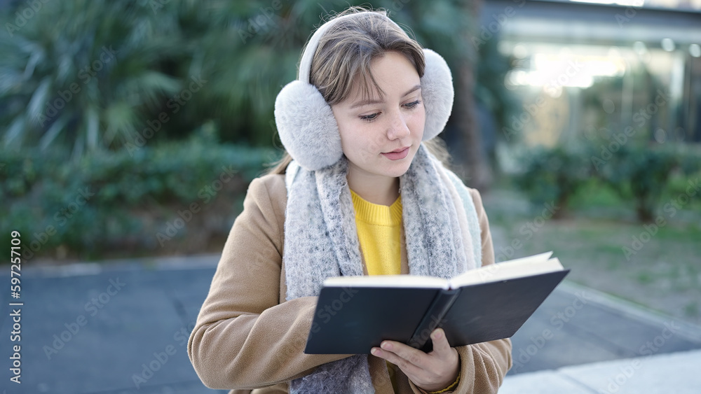 Fototapeta premium Young blonde woman reading book sitting on bench at park