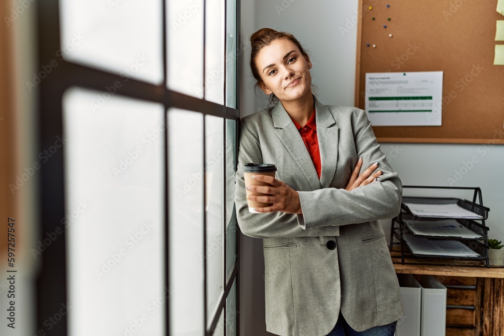 Young woman business worker drinking coffee standing with arms crossed gesture at office