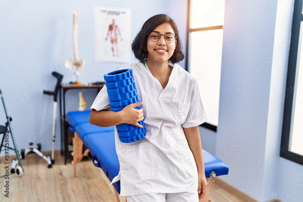 Young latin woman wearing physiotherapist uniform holding foam roller at physiotherapy clinic Young latin woman wearing physiotherapist uniform holding foam roller at physiotherapy clinic