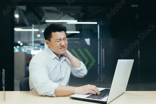 An office worker of Asian origin is working on a laptop and holding his neck. Suffering and writhing in pain, he needs medical help.