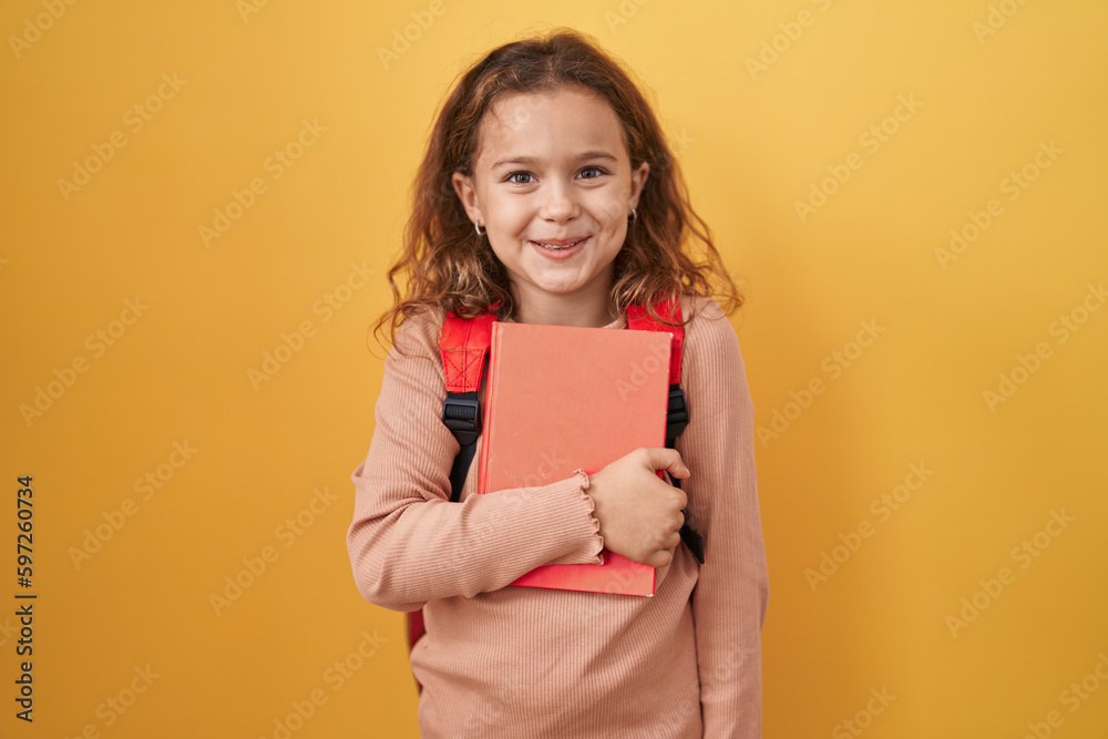Little caucasian girl wearing student backpack and holding book looking positive and happy standing and smiling with a confident smile showing teeth Little caucasian girl wearing student backpack and holding book looking positive and happy standing and smiling with a confident smile showing teeth