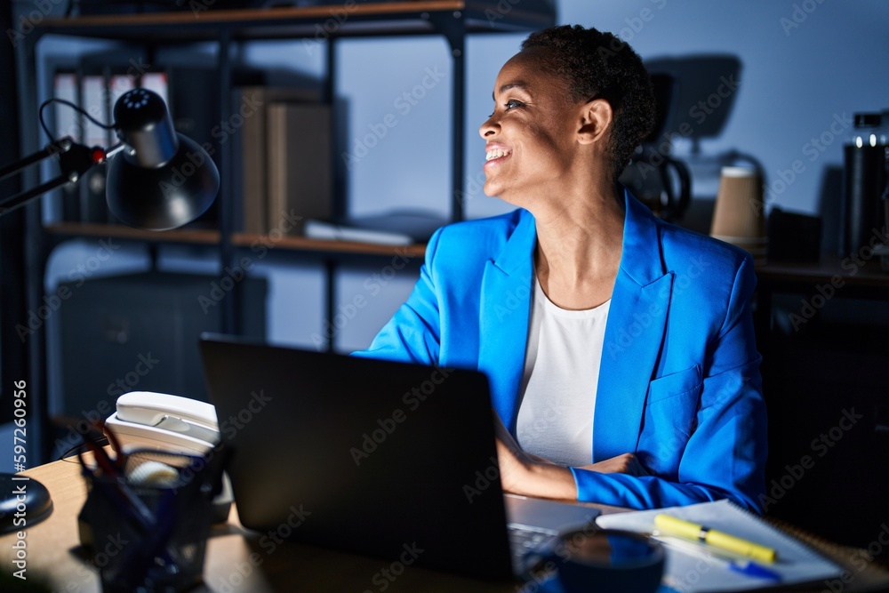 Beautiful african american woman working at the office at night looking away to side with smile on face, natural expression. laughing confident.