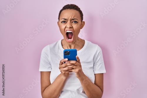 Beautiful african american woman using smartphone angry and mad screaming frustrated and furious, shouting with anger. rage and aggressive concept.