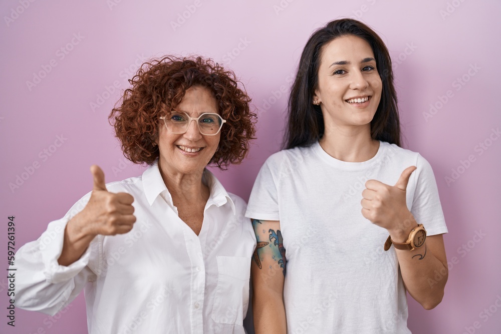 Hispanic mother and daughter wearing casual white t shirt over pink background doing happy thumbs up gesture with hand. approving expression looking at the camera showing success. Hispanic mother and daughter wearing casual white t shirt over pink background doing happy thumbs up gesture with hand. approving expression looking at the camera showing success.