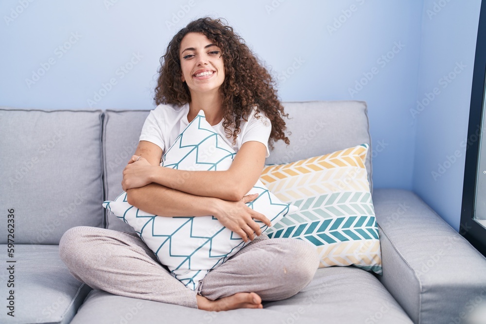 Hispanic woman with curly hair sitting on the sofa at home smiling with a happy and cool smile on face. showing teeth. Hispanic woman with curly hair sitting on the sofa at home smiling with a happy and cool smile on face. showing teeth.
