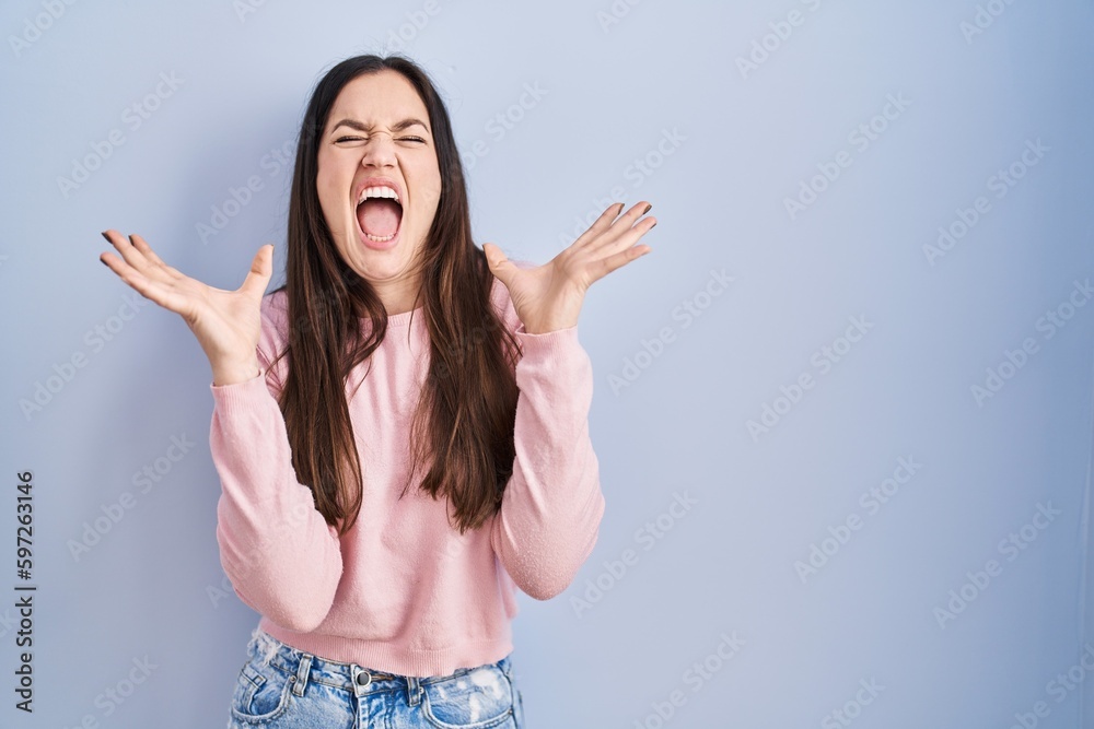 Young brunette woman standing over blue background crazy and mad shouting and yelling with aggressive expression and arms raised. frustration concept.