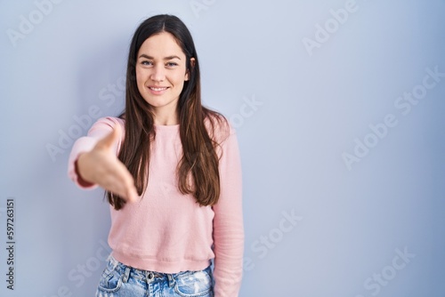 Young brunette woman standing over blue background smiling friendly offering handshake as greeting and welcoming. successful business. © Krakenimages.com
