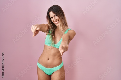Young hispanic woman wearing bikini over pink background pointing to you and the camera with fingers, smiling positive and cheerful