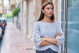 Young beautiful hispanic woman standing with serious expression and arms crossed gesture at street