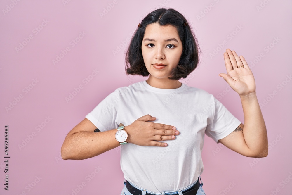 Young hispanic woman wearing casual white t shirt over pink background swearing with hand on chest and open palm, making a loyalty promise oath Young hispanic woman wearing casual white t shirt over pink background swearing with hand on chest and open palm, making a loyalty promise oath