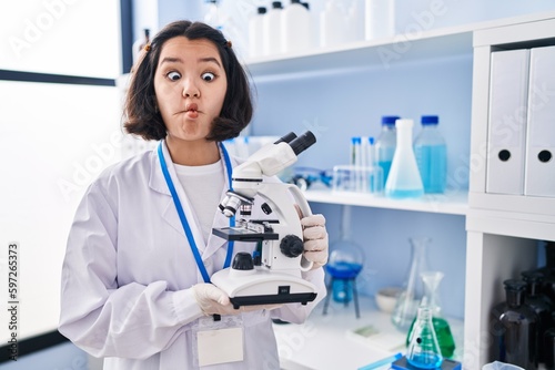 Young hispanic woman working at scientist laboratory holding microscope making fish face with mouth and squinting eyes  crazy and comical.
