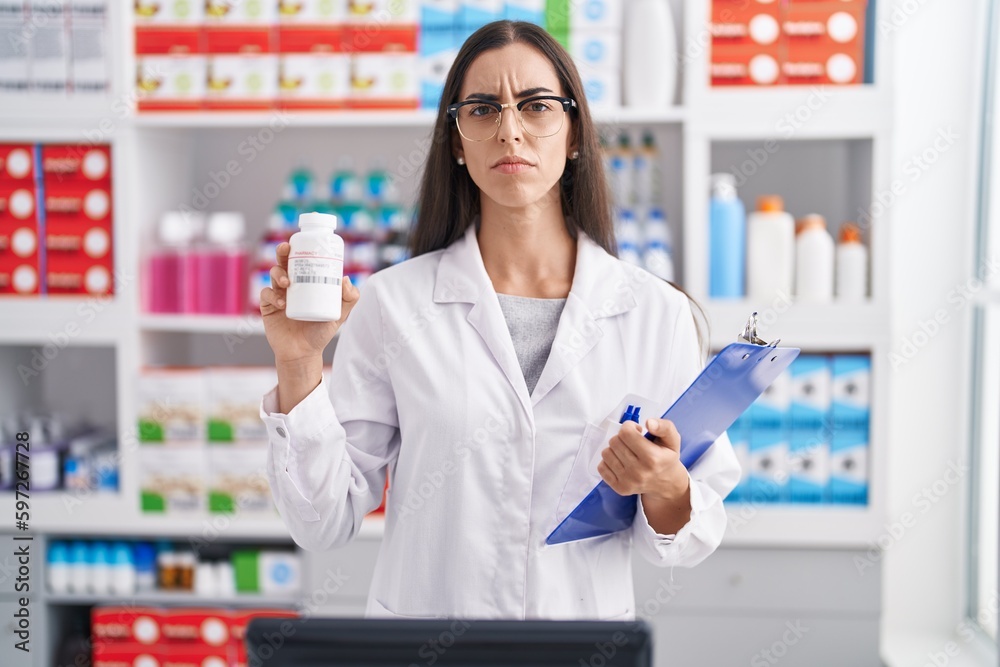 Young brunette woman working at pharmacy drugstore holding pills skeptic and nervous, frowning upset because of problem. negative person.