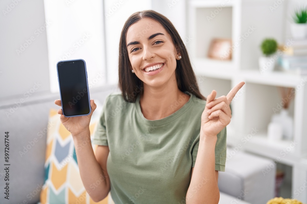 Young brunette woman holding smartphone showing blank screen smiling happy pointing with hand and finger to the side
