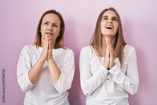 Middle age mother and young daughter standing over pink background begging and praying with hands together with hope expression on face very emotional and worried. begging.