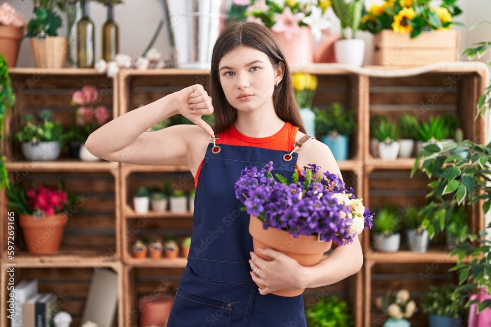 Young caucasian woman working at florist shop holding pot with flowers with angry face, negative sign showing dislike with thumbs down, rejection concept