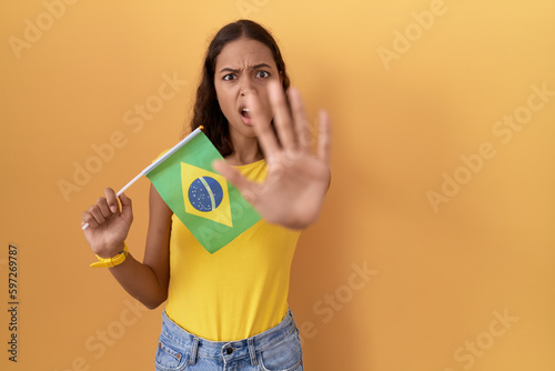 Young hispanic woman holding brazil flag doing stop gesture with hands palms, angry and frustration expression