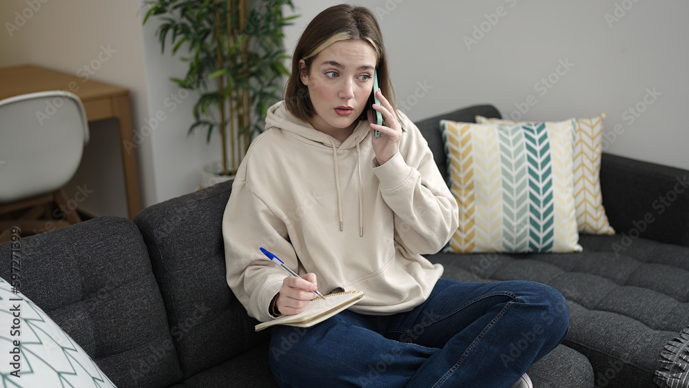 Young blonde woman writing on notebook talking on smartphone at home