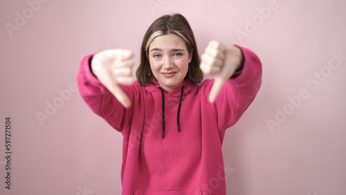 Young blonde woman doing negative gesture with thumbs down over isolated pink background